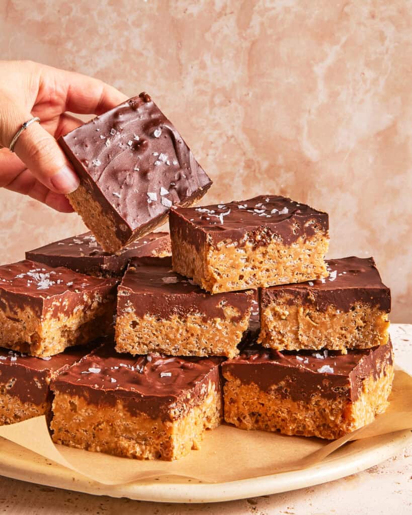 A hand picks up a chocolate-topped dessert bar from a plate stacked with similar bars. The bars have a crispy base and are sprinkled with flaky sea salt.