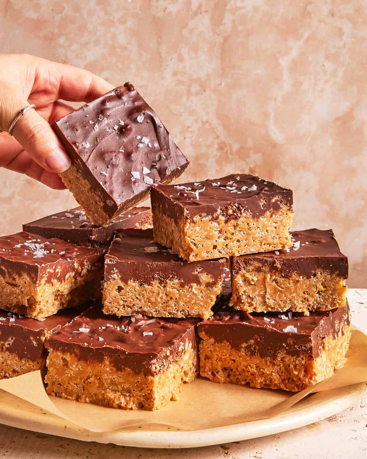 A hand picks up a chocolate-topped dessert bar from a plate stacked with similar bars. The bars have a crispy base and are sprinkled with flaky sea salt.