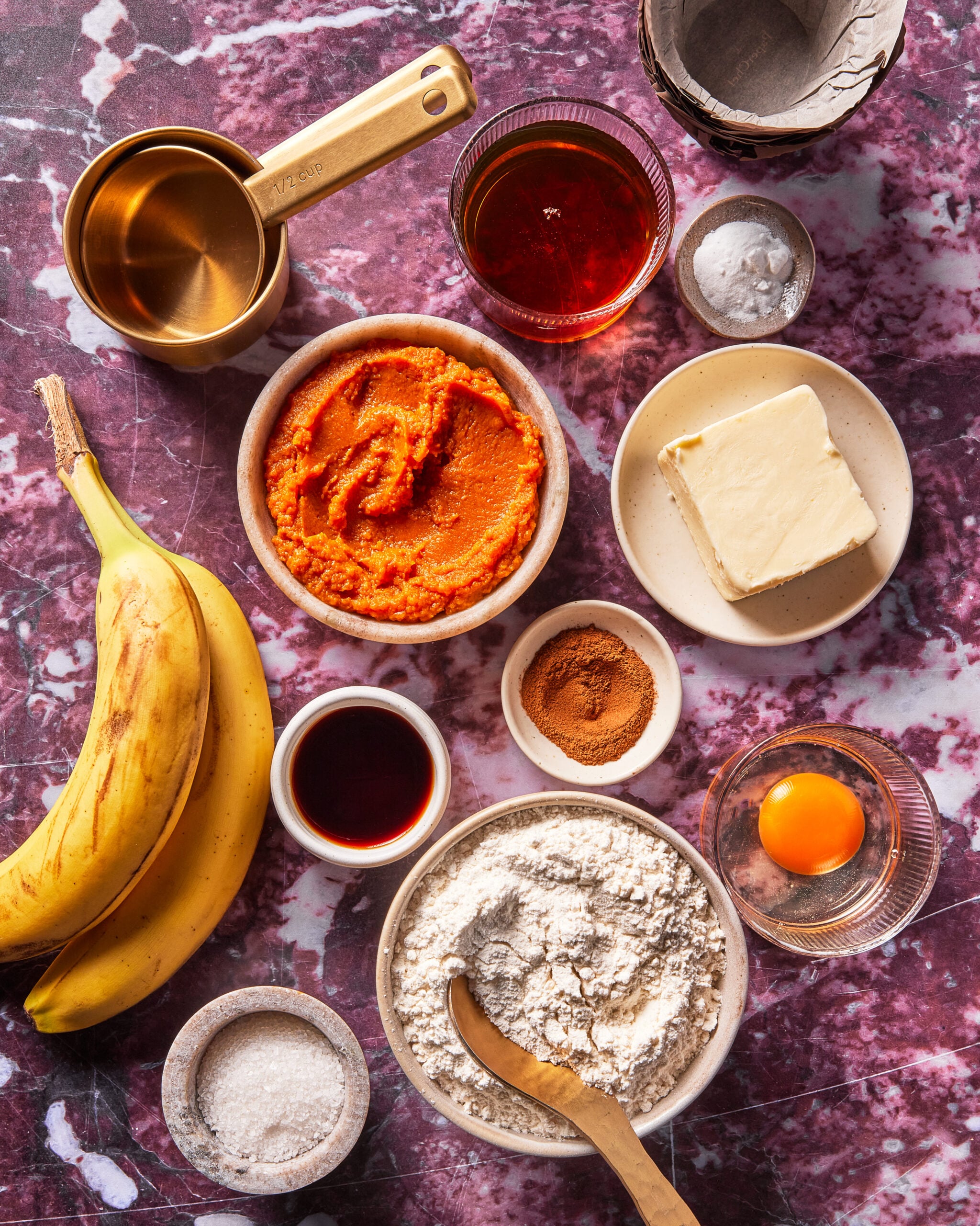 Baking ingredients on a marble surface: bananas, pumpkin puree, butter, flour, egg yolk, sugar, cinnamon, vanilla, baking powder, liquid sweetener, and measuring cup.