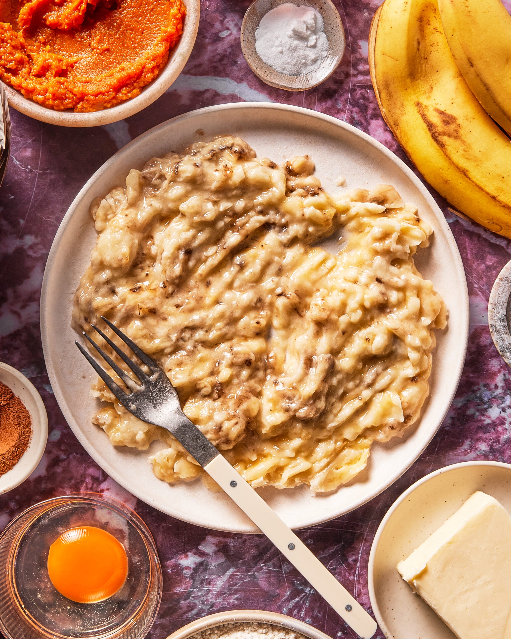 A plate of mashed bananas with a fork, surrounded by baking ingredients like butter, an egg, cinnamon, baking soda, pumpkin puree, and whole bananas on a marbled surface.
