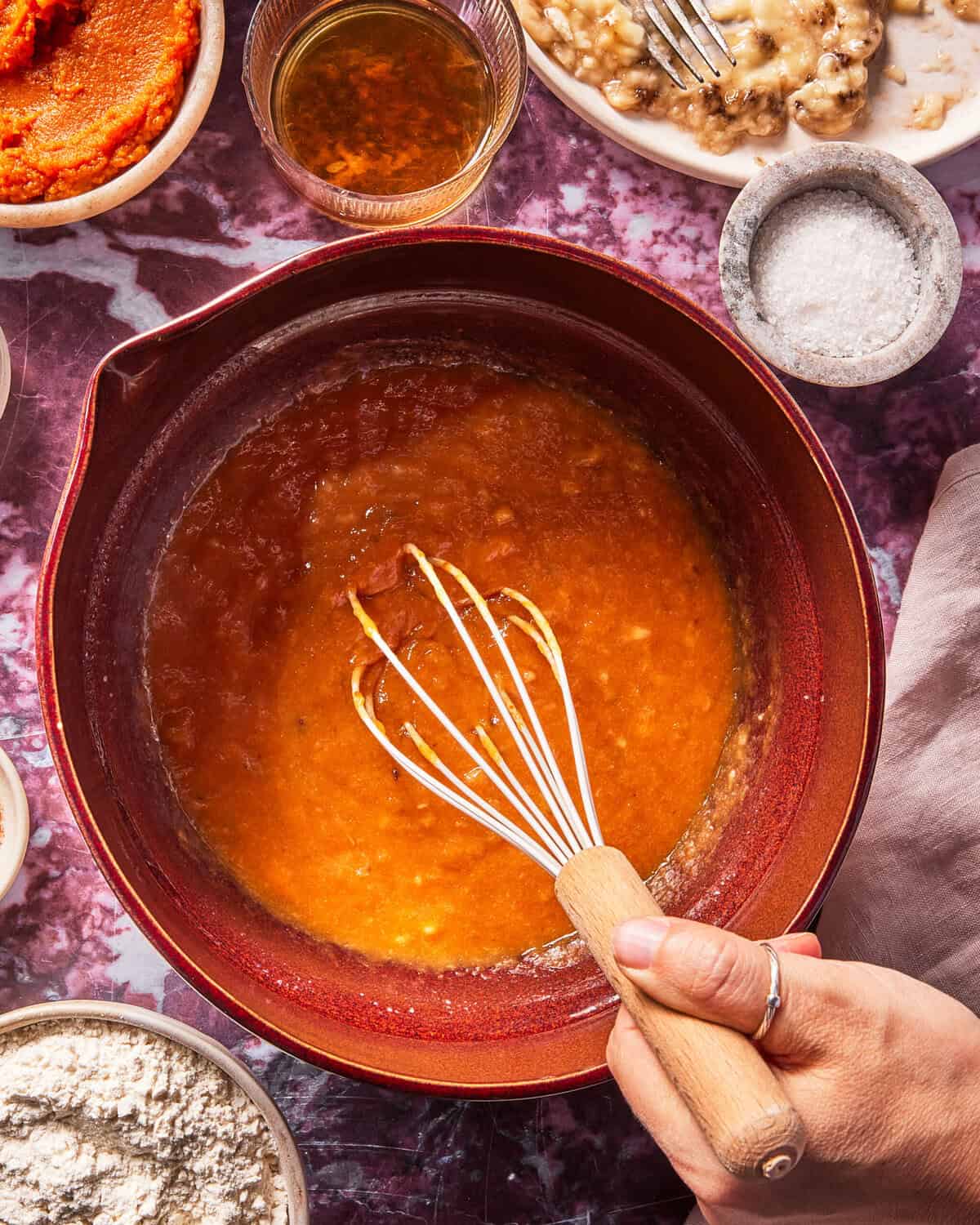 A hand whisking orange batter in a red mixing bowl, surrounded by bowls of flour, salt, oil, and pumpkin puree on a purple marbled surface.