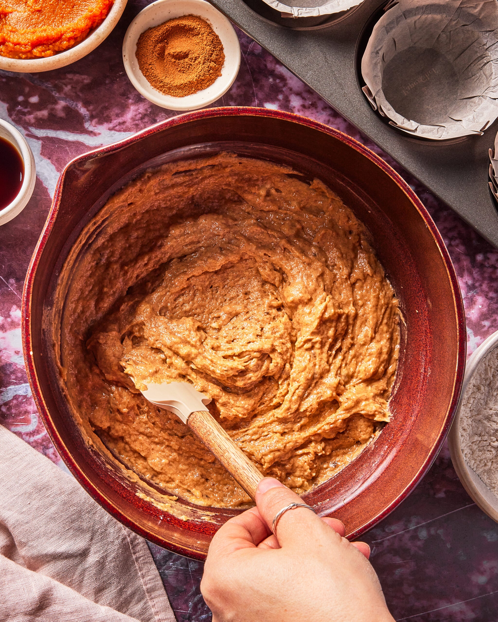 A hand stirs thick brown batter in a large red bowl with a spatula. Surrounding the bowl are small dishes of spices, vanilla, and flour on a purple marbled countertop.