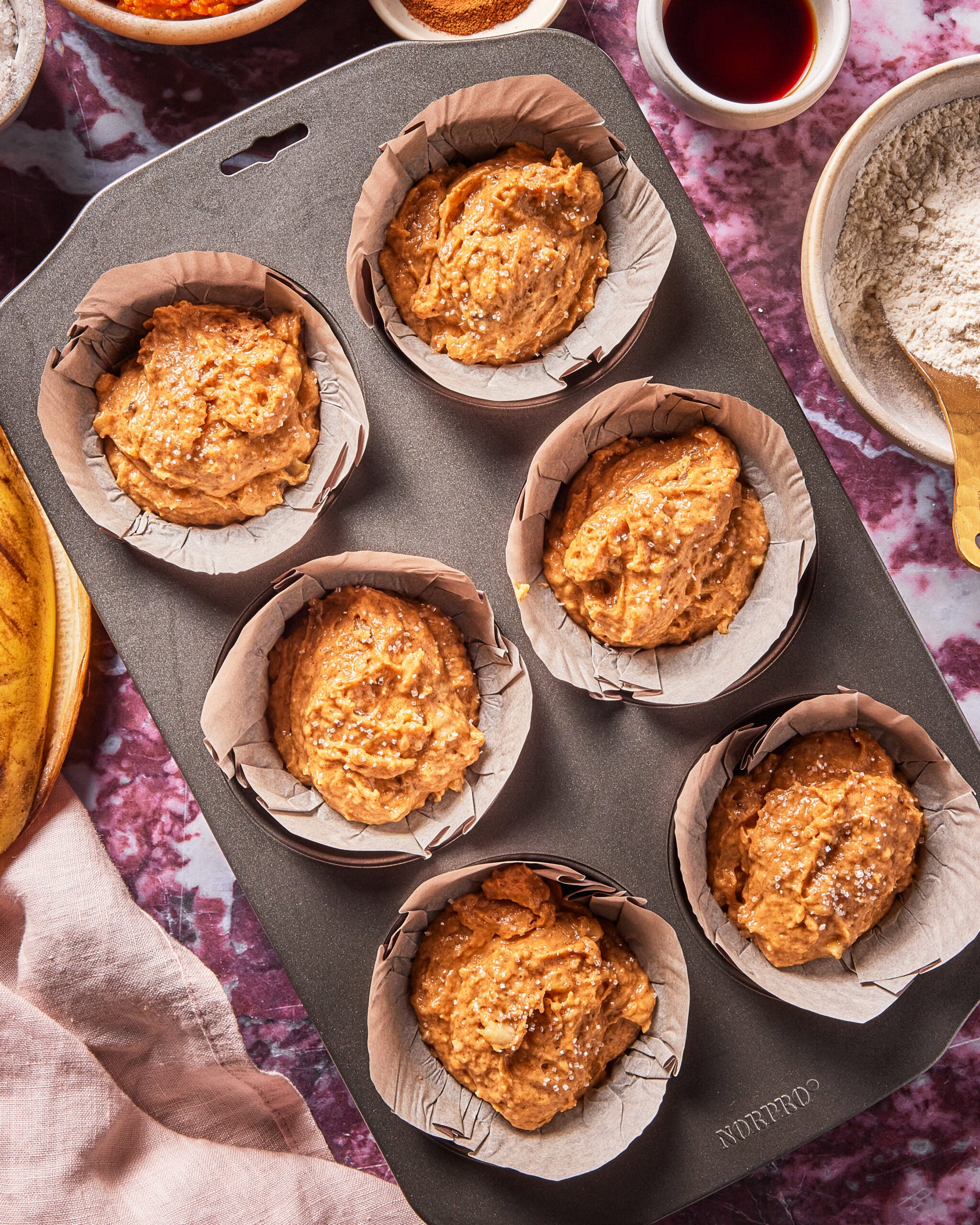 A muffin tray with six sections, each lined with parchment paper and filled with muffin batter, sits on a purple countertop surrounded by bowls of ingredients.