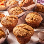 Pumpkin muffins with golden tops are arranged on a marble board, some in parchment wrappers. Bowls of flour, cinnamon, and pumpkin puree are in the background.