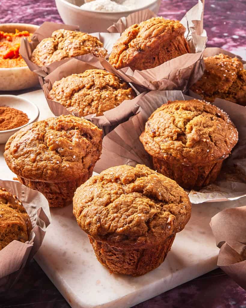 Pumpkin muffins with golden tops are arranged on a marble board, some in parchment wrappers. Bowls of flour, cinnamon, and pumpkin puree are in the background.