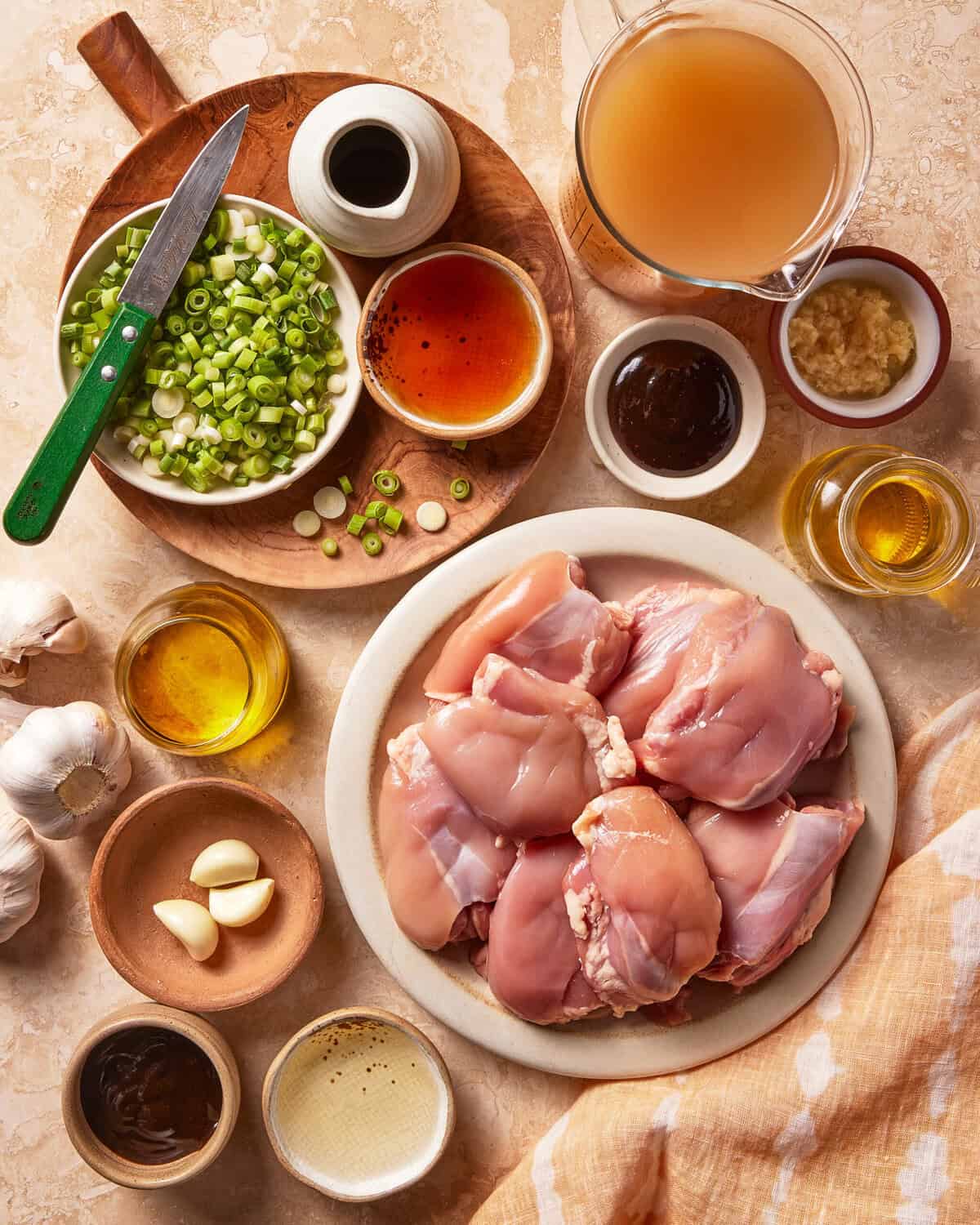 Raw chicken thighs on a plate surrounded by chopped green onions, garlic, ginger, sauces, oil, broth, and a knife on a cutting board, all arranged on a beige countertop.