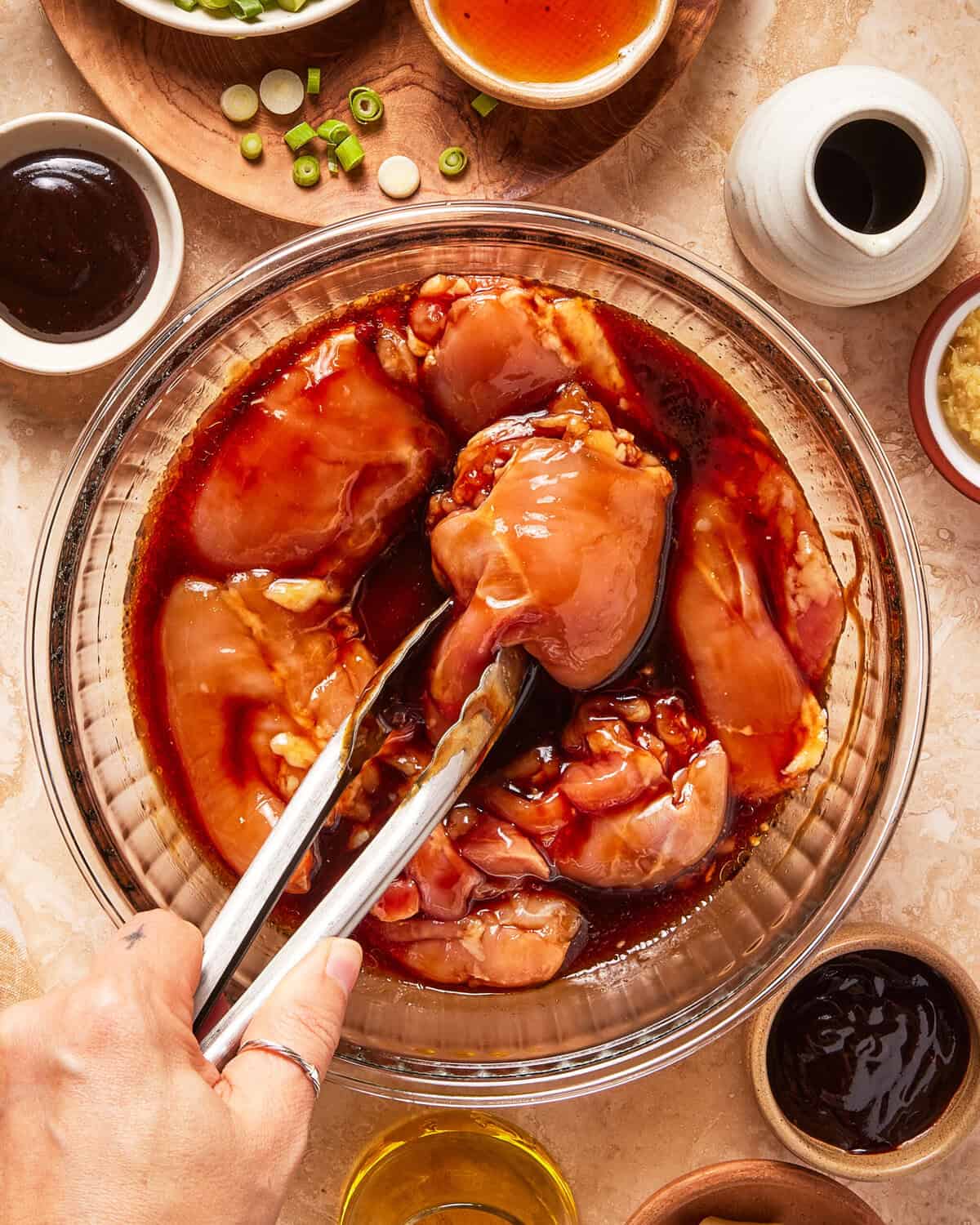 A hand uses tongs to mix raw chicken thighs in a glass bowl with marinade. Various bowls of sauces, oil, green onions, and minced garlic are arranged around the bowl on a beige countertop.