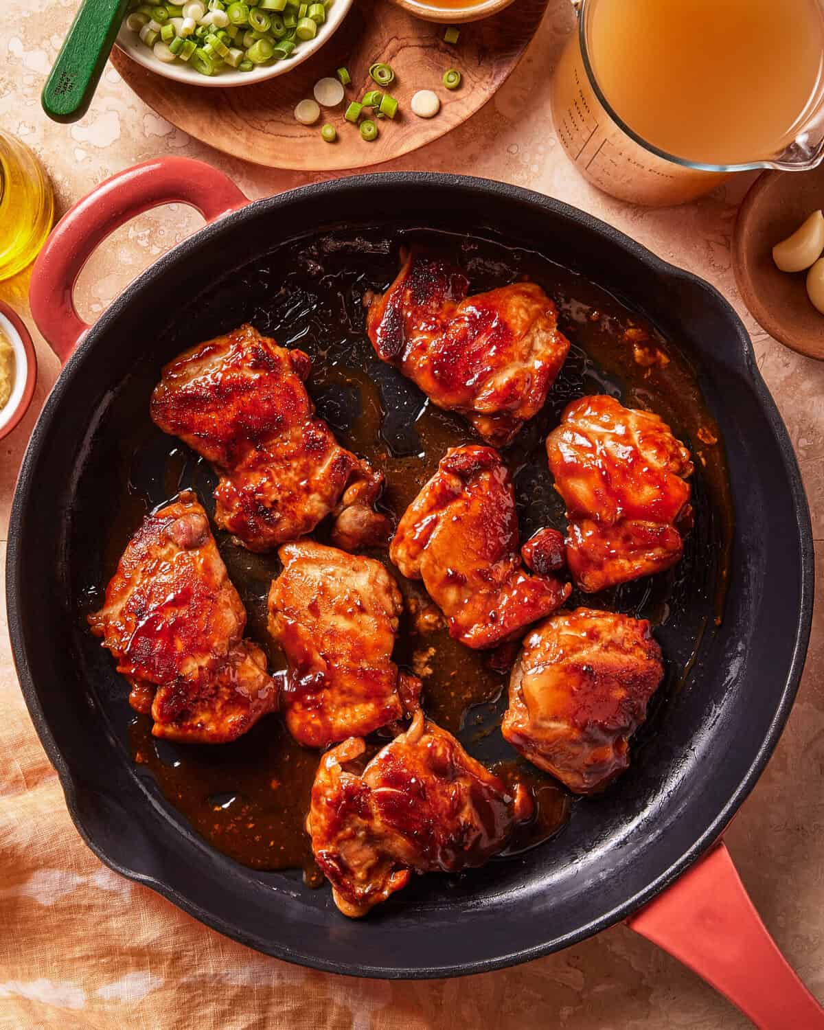 Seven glazed chicken thighs cook in a round black skillet with red handles. Surrounding the pan are green onions, broth, garlic, and other cooking ingredients on a beige surface.