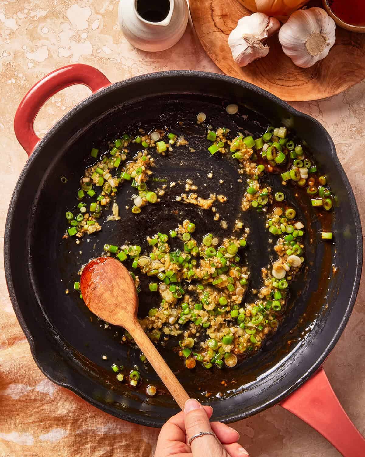 A hand stirs chopped green onions and minced garlic in a black skillet with a wooden spoon. Garlic bulbs, a sauce jar, and a bowl sit nearby on a beige countertop.