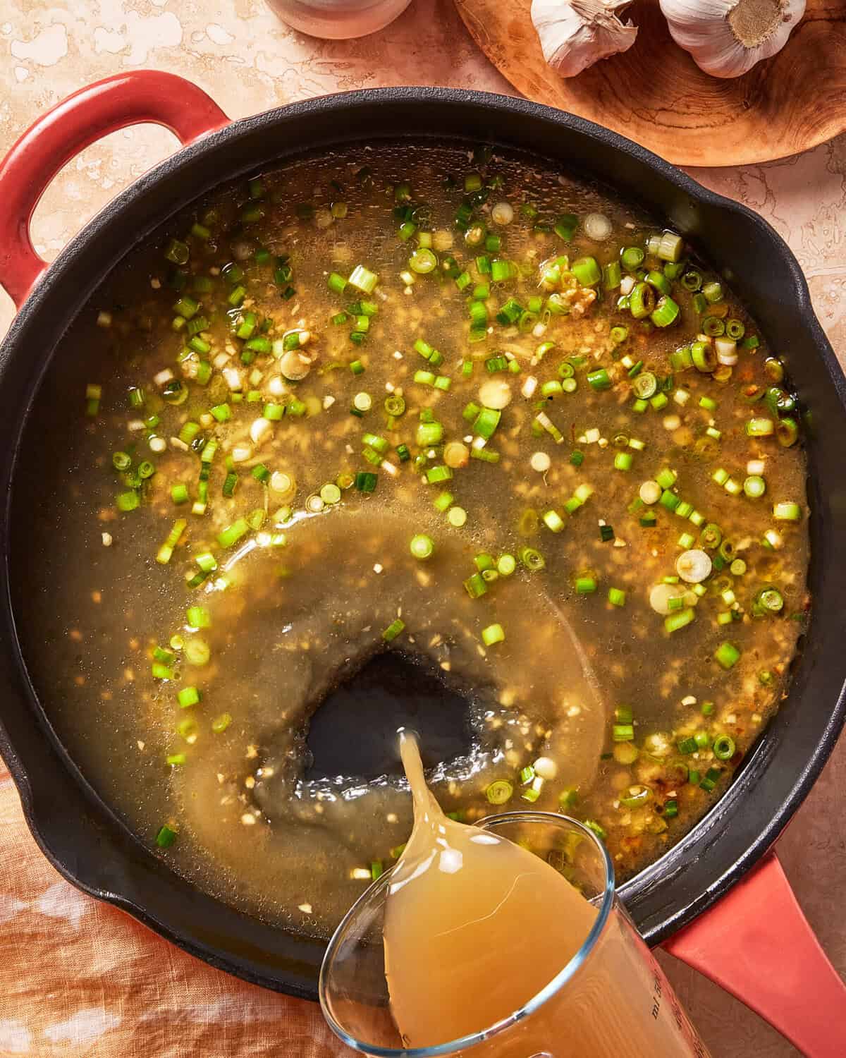 A hand pours broth from a measuring cup into a skillet filled with chopped green onions and spices, creating a swirl in the liquid. Garlic bulbs and a wooden bowl sit nearby on the table.