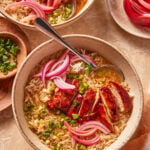 A bowl of rice soup topped with sliced glazed chicken, pickled red onions, and chopped green onions. A spoon rests in the bowl, with additional garnishes and another bowl in the background.