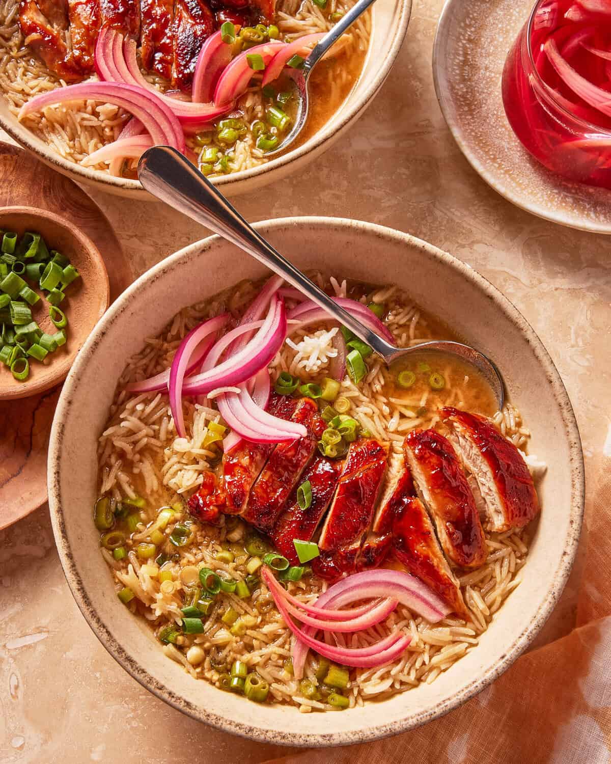 A bowl of rice soup topped with sliced glazed chicken, pickled red onions, and chopped green onions. A spoon rests in the bowl, with additional garnishes and another bowl in the background.