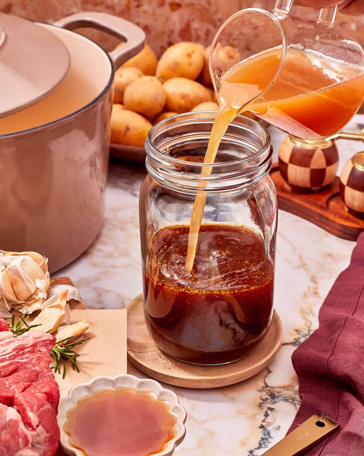 Broth being poured from a glass pitcher into a glass jar containing a dark sauce, with ingredients including garlic and raw meat nearby.