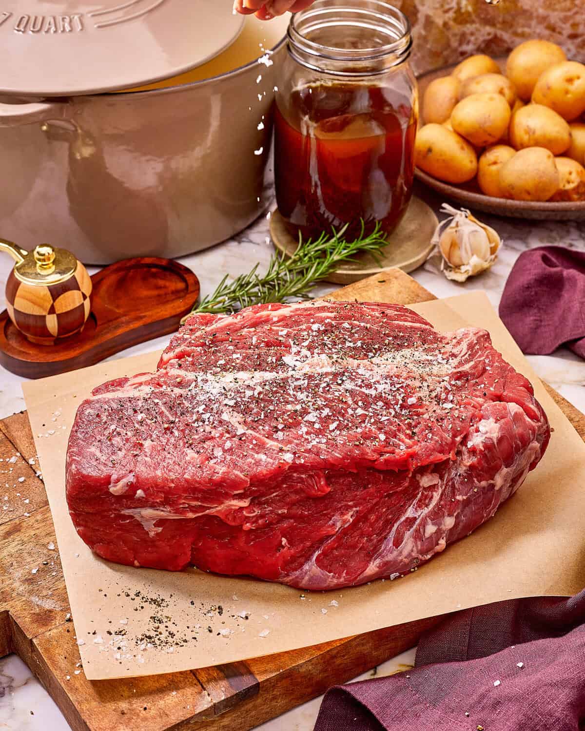 A raw beef roast on a wooden cutting board, being seasoned with salt and pepper.