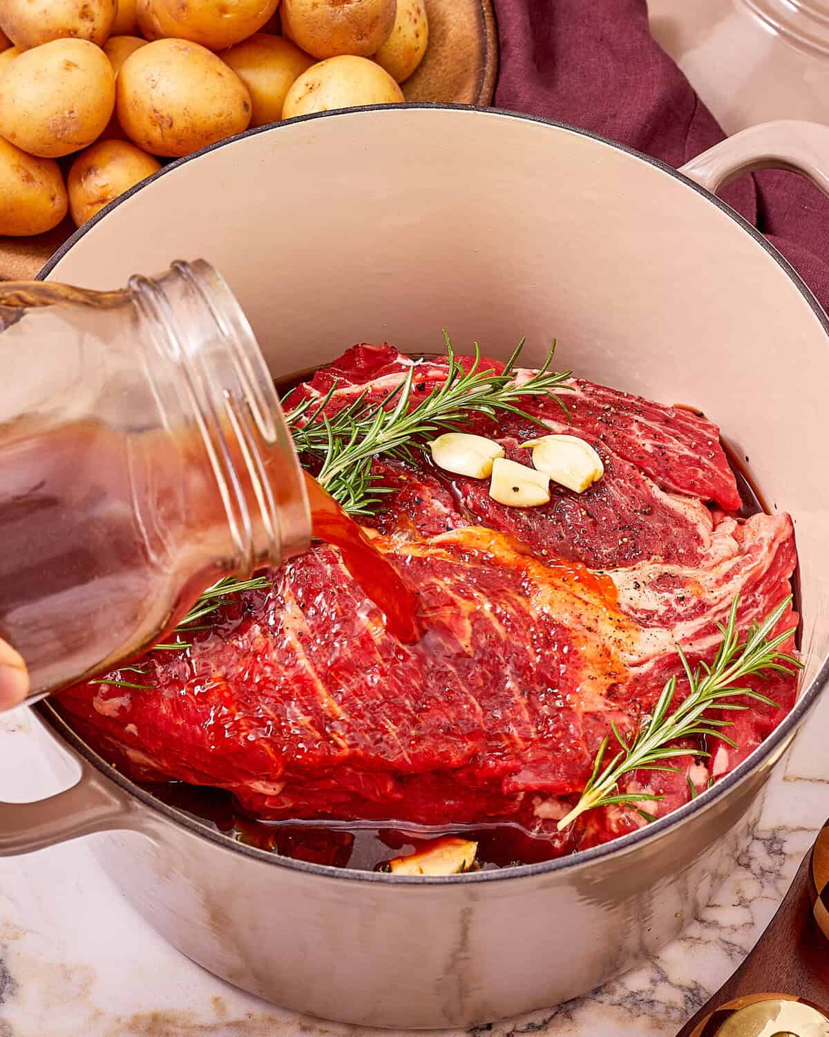 Liquid being poured from a jar over a large piece of raw beef in a Dutch oven, topped with garlic cloves and rosemary sprigs.