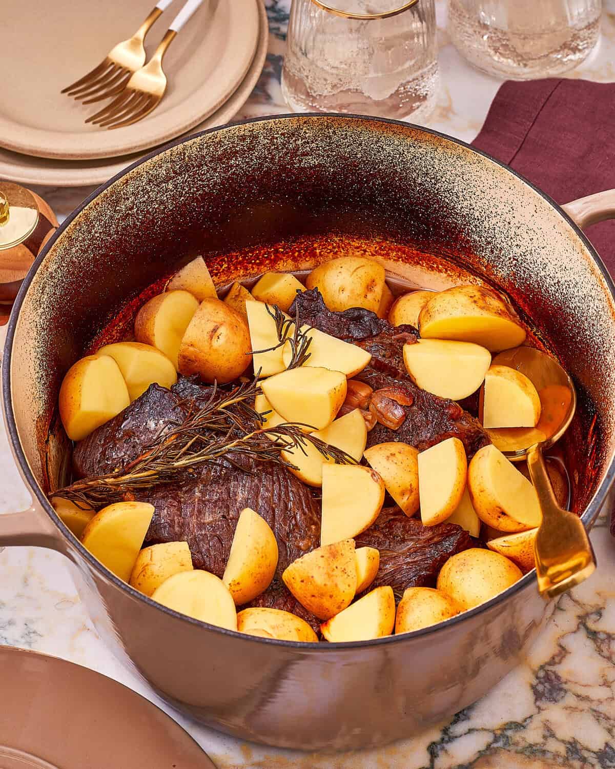A Dutch oven filled with a pot roast, chunks of potato, and a sprig of rosemary, with a serving spoon resting inside.