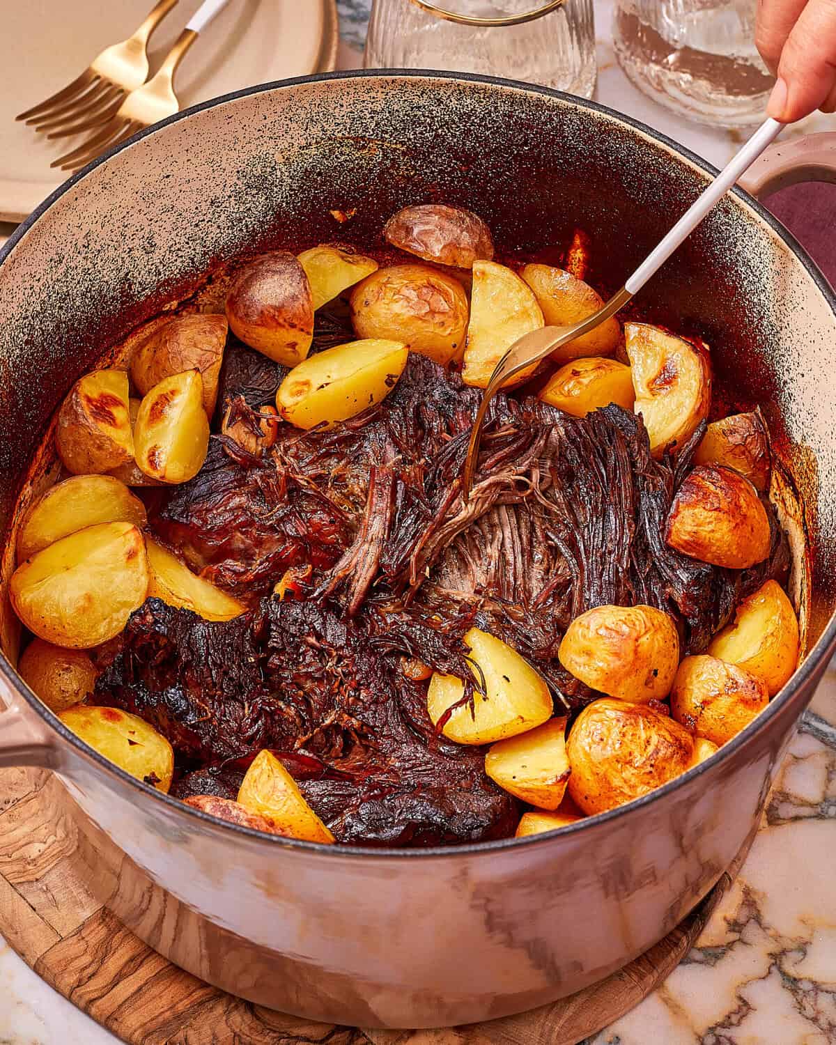 A fork pulls apart a tender pot roast inside a Dutch oven surrounded by roasted potatoes.