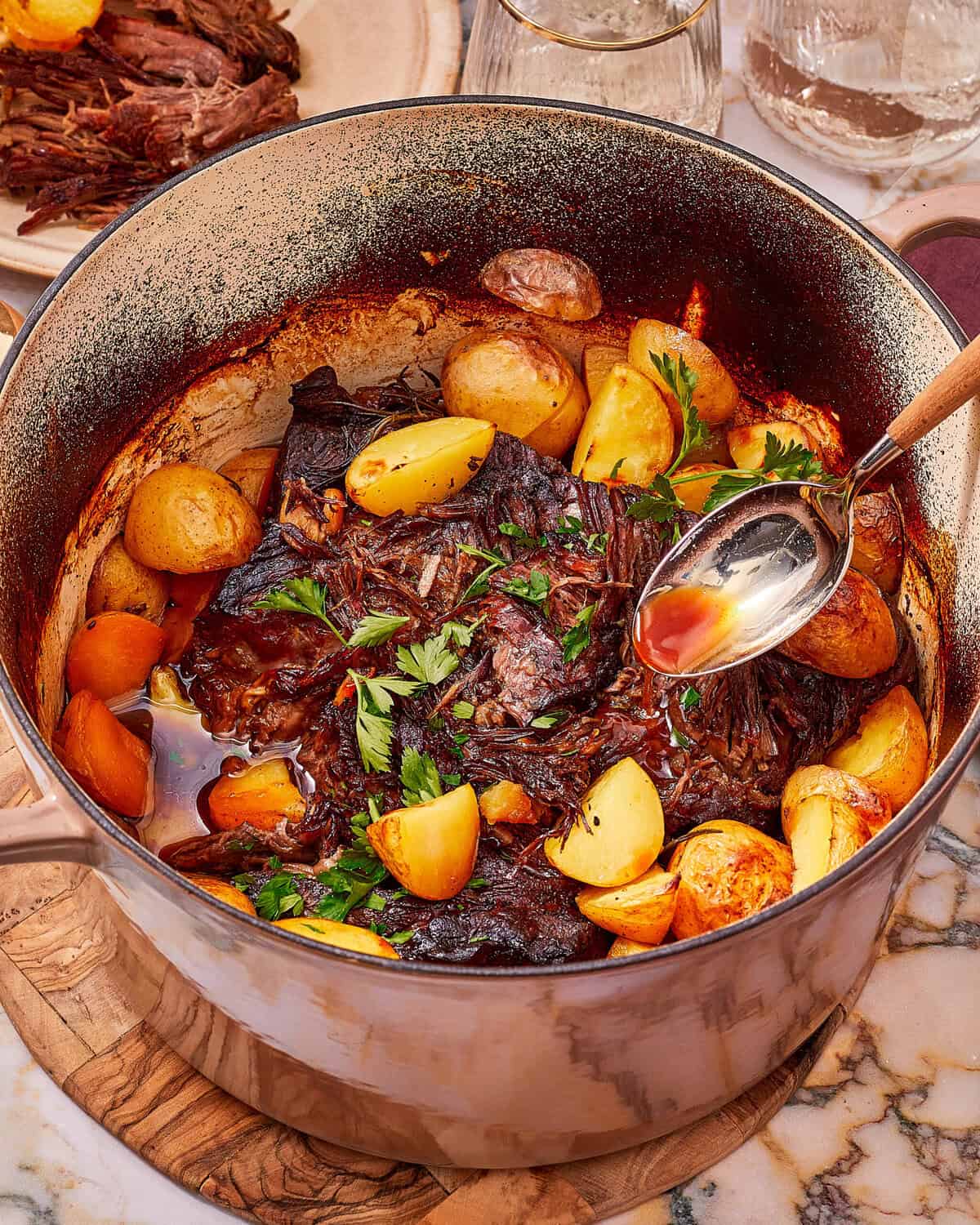 A Dutch oven filled with braised pot roast and roasted potatoes garnished with fresh parsley, with a spoon lifting juices from the pot.