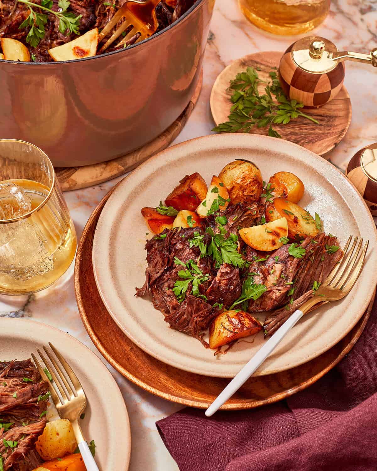 A plate of beef pot roast with roasted potatoes, garnished with fresh parsley, next to a glass of iced tea and a large Dutch oven.