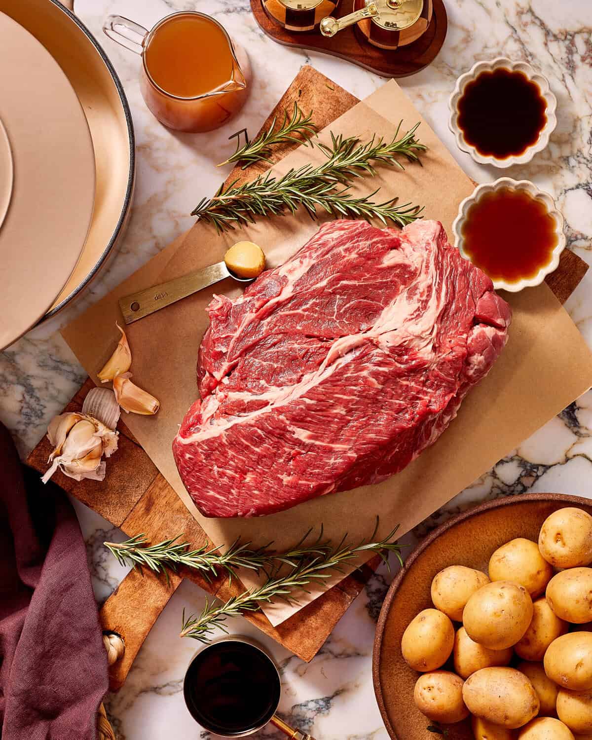 A raw beef chuck roast on parchment paper, surrounded by rosemary sprigs, garlic cloves, bowls of sauce, and a bowl of baby potatoes.
