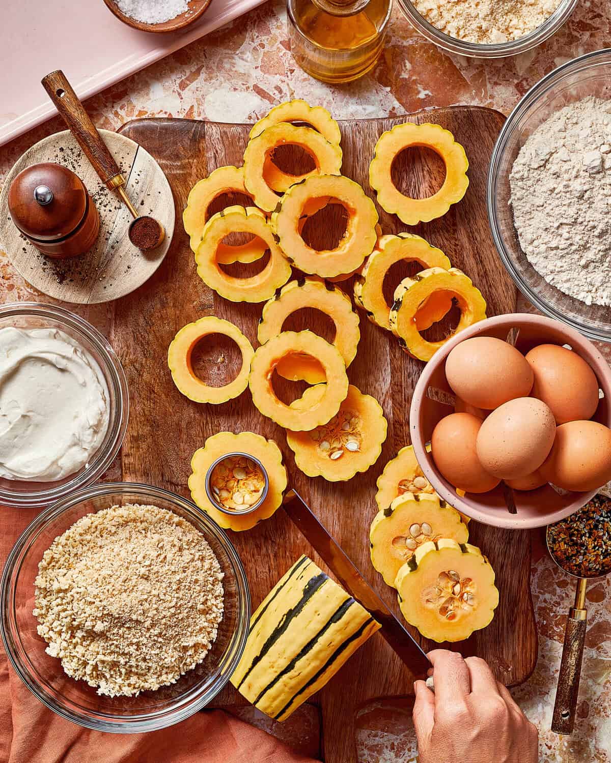 A hand uses a knife to slice a striped acorn squash on a wooden board surrounded by bowls of breadcrumbs, eggs, flour, and cream.