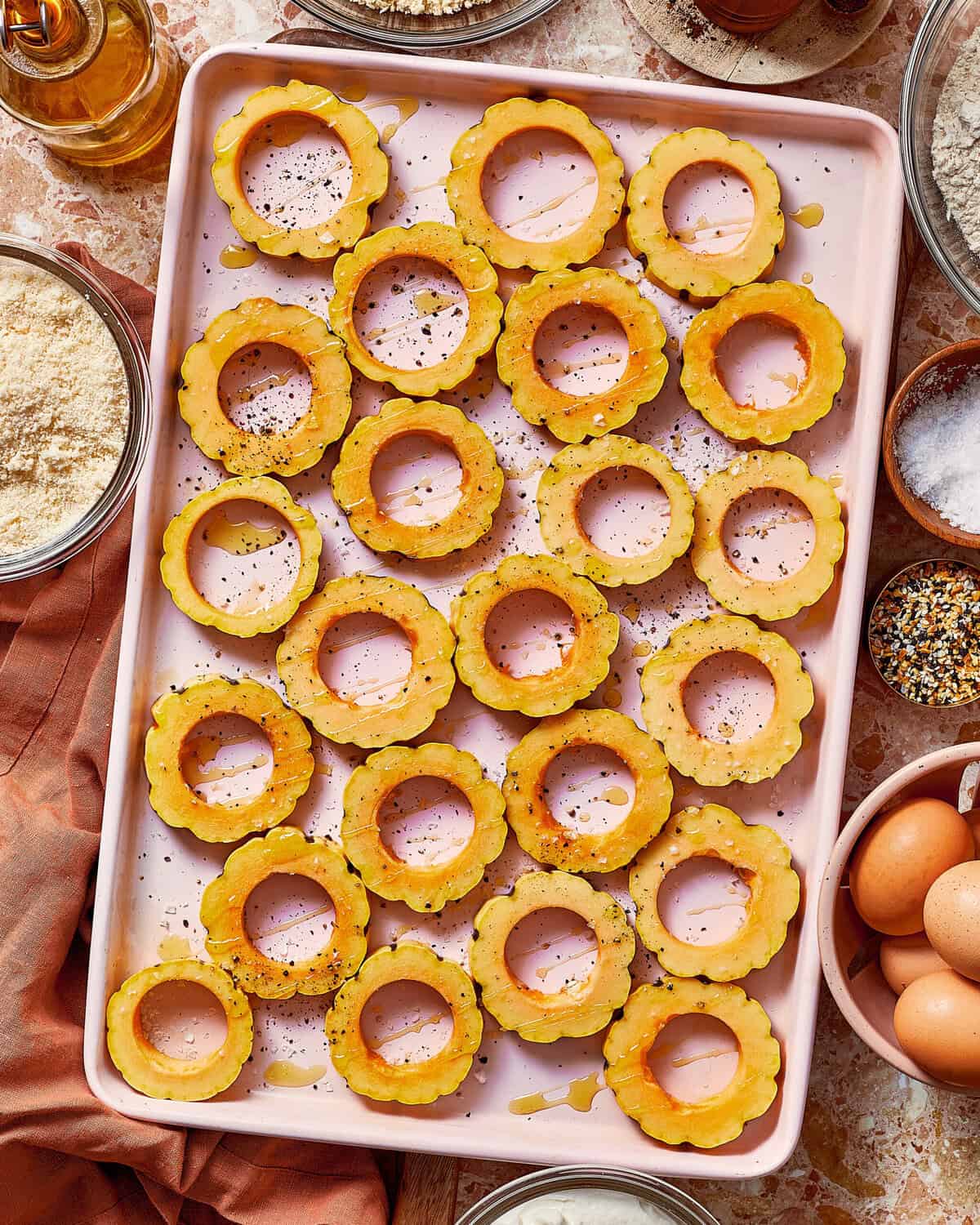 A pink baking sheet filled with raw, circular slices of yellow delicata squash, seasoned with pepper and drizzled with oil.