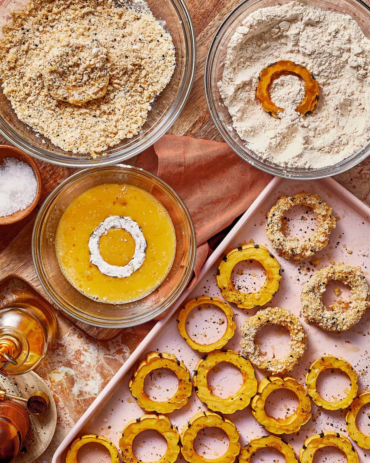 Bowls of flour, egg wash, and breadcrumbs used to coat slices of squash on a baking sheet.