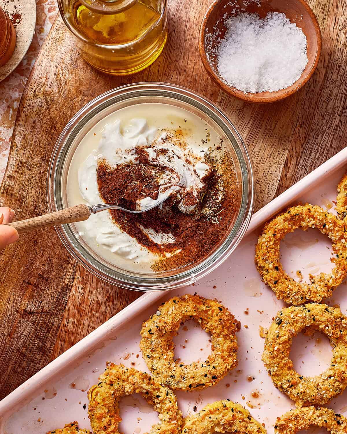 A hand stirs red spice into a glass bowl of white sauce, next to a pink tray of baked onion rings.