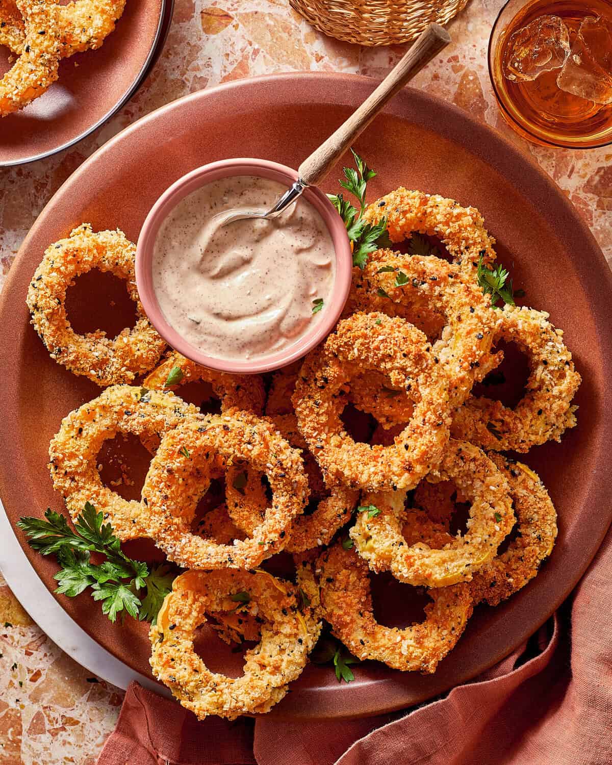 A plate of breaded onion rings garnished with parsley, served with a small bowl of dipping sauce and a wooden spoon.