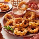 A hand dips a crispy breaded onion ring into a bowl of creamy dipping sauce on a platter of golden-brown onion rings.