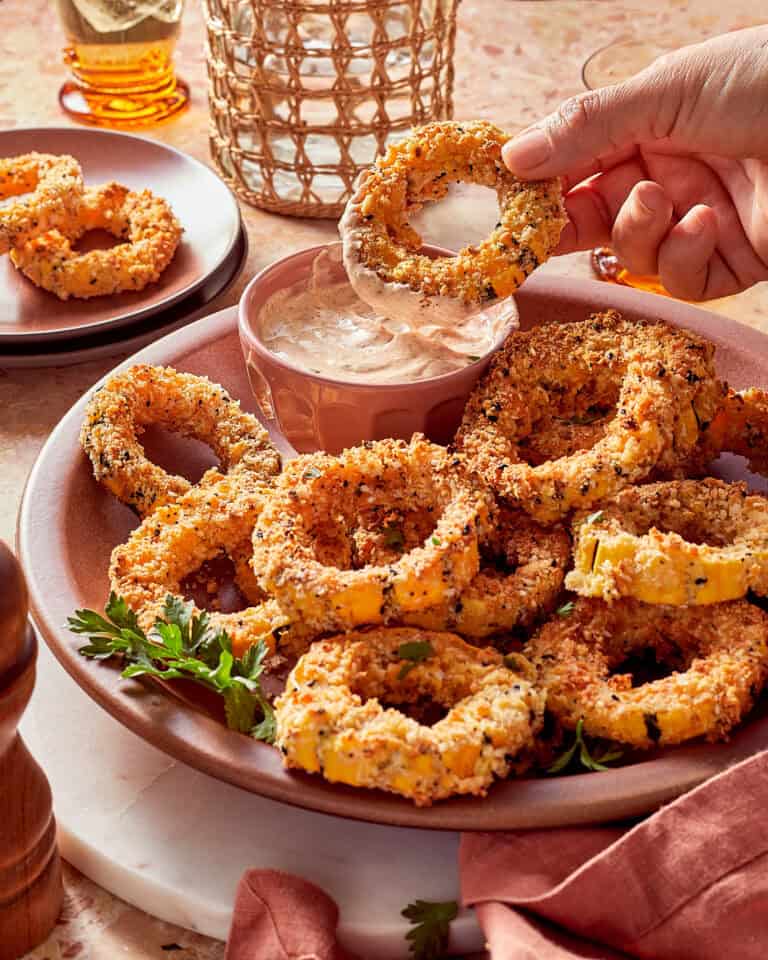 A hand dips a crispy breaded onion ring into a bowl of creamy dipping sauce on a platter of golden-brown onion rings.
