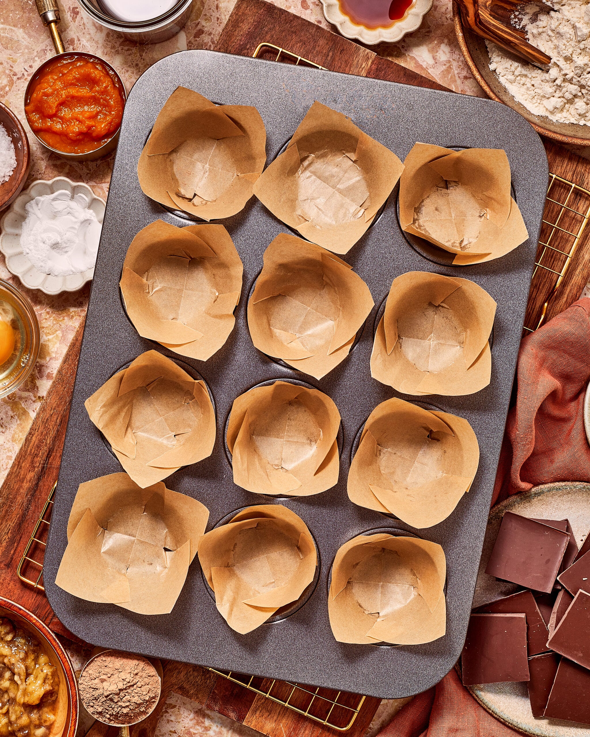 A muffin tin lined with parchment paper liners, surrounded by bowls of ingredients including pumpkin puree, flour, cocoa powder, and dark chocolate squares.