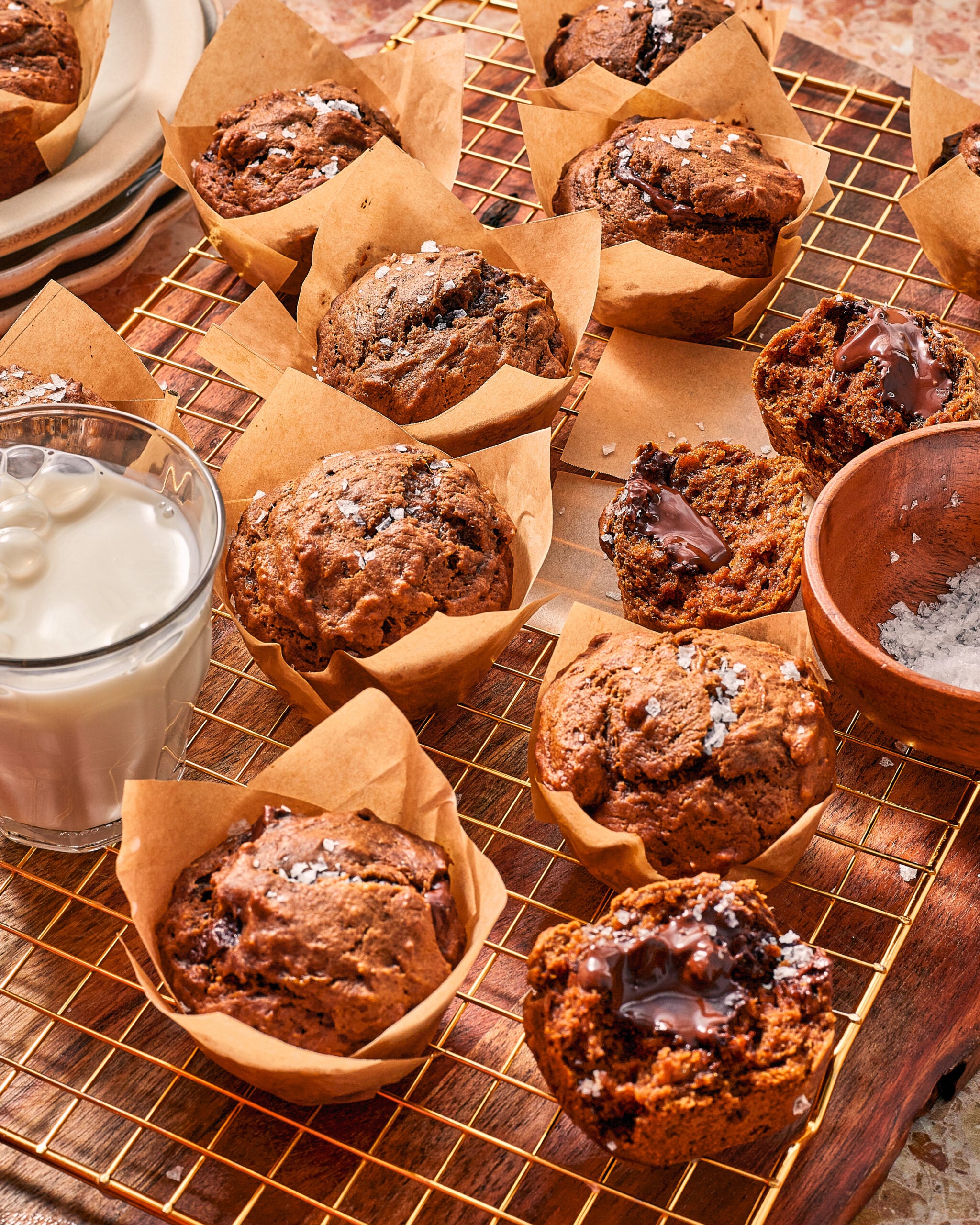 A wire cooling rack filled with chocolate muffins topped with sea salt, including some broken open to reveal melted chocolate centers, next to a glass of milk and a small bowl of salt.