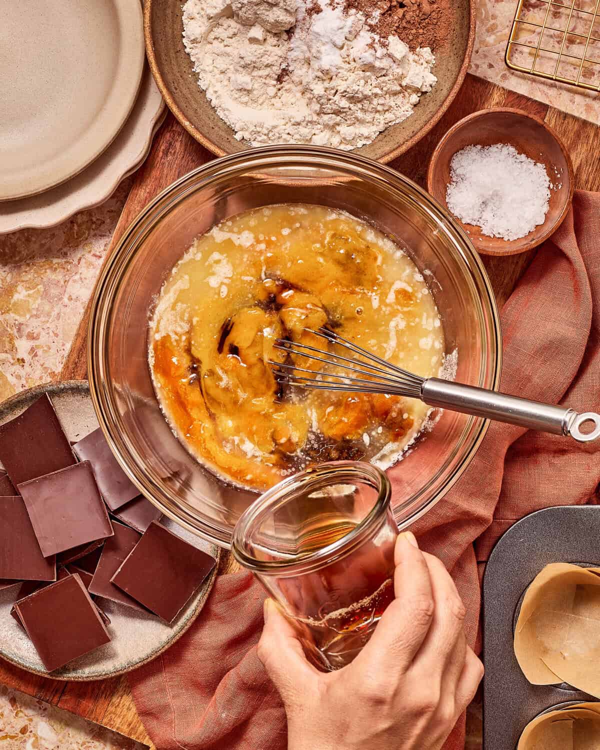 A hand pours liquid from a small glass into a mixing bowl containing whisked eggs, oil, and vanilla, with bowls of flour and chocolate squares nearby.
