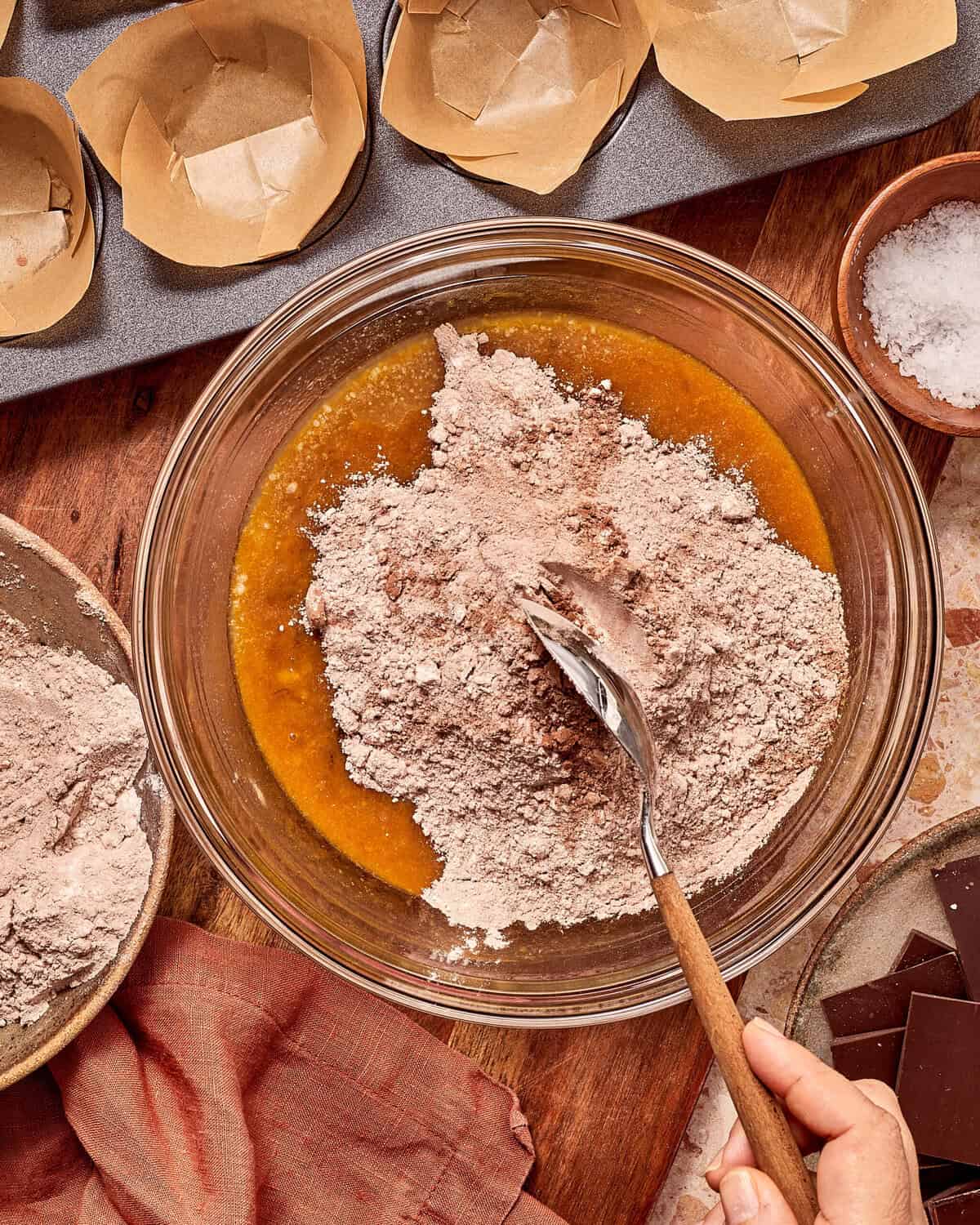 A hand holds a spoon mixing a pile of dry chocolate powder into a golden liquid base in a glass bowl, with muffin liners and chocolate squares nearby.