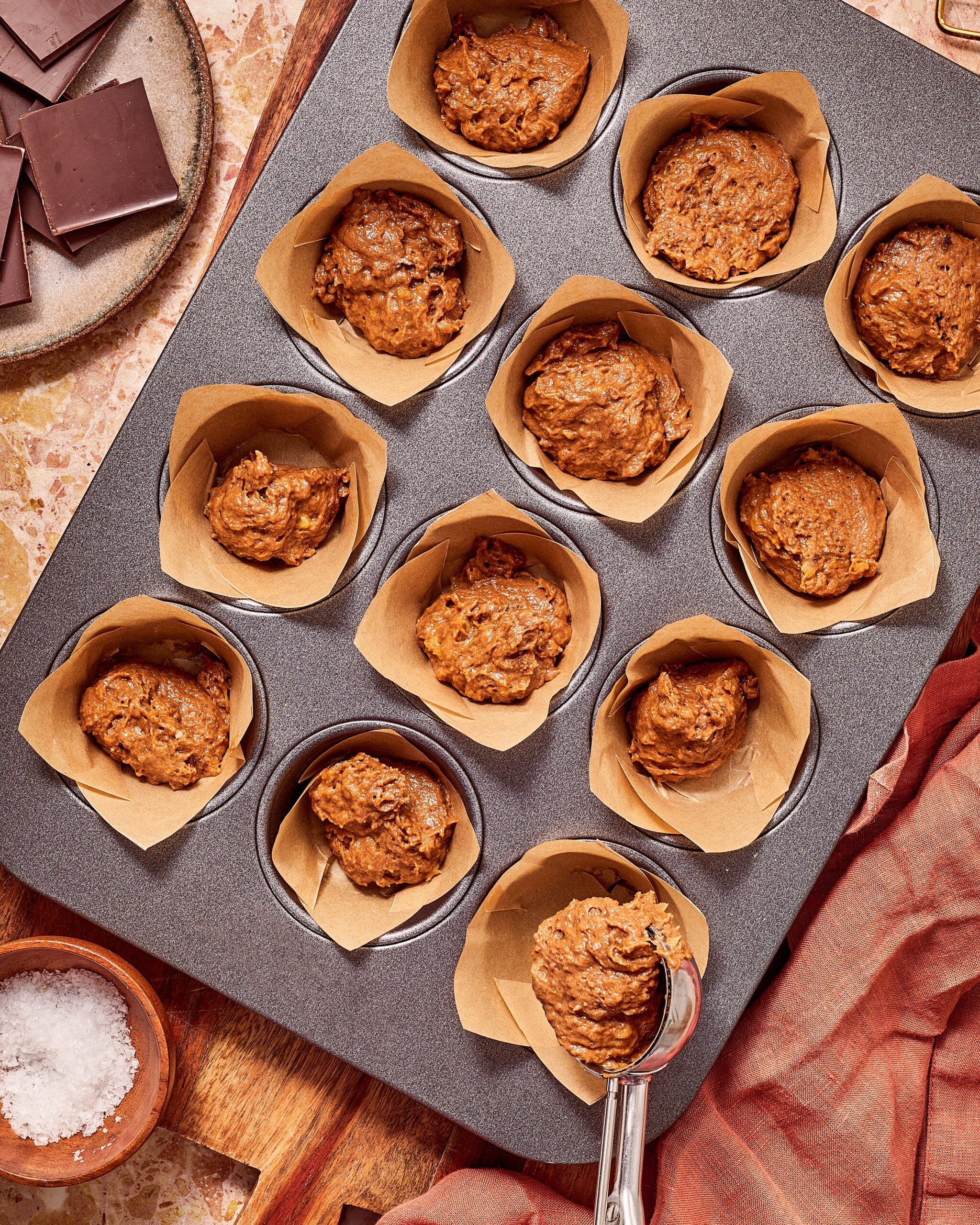 A muffin tin with paper liners filled with scoops of chocolate batter, with a scoop held over one section, next to a small bowl of sea salt and a plate of chocolate bars.