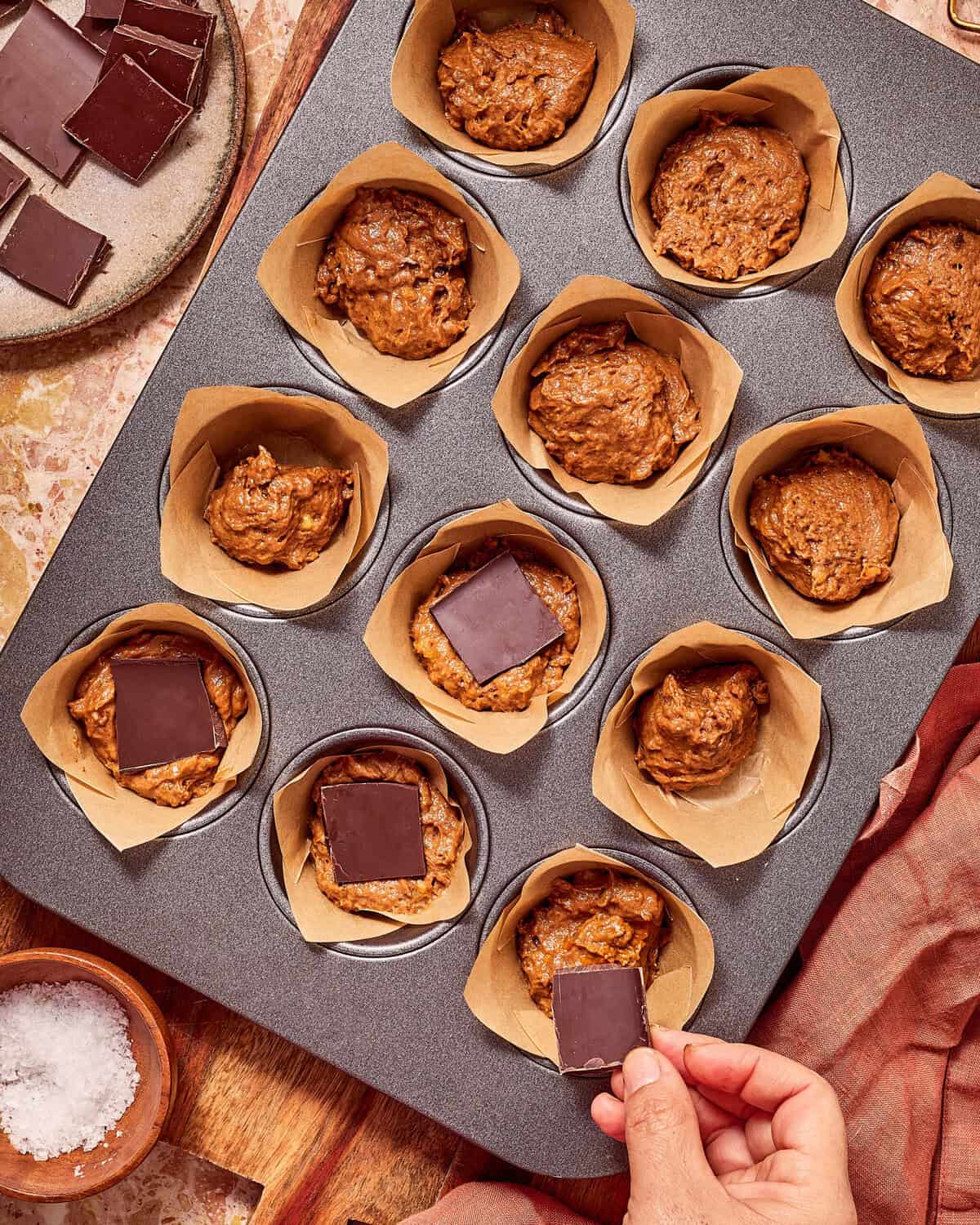 A hand places a square of dark chocolate onto muffin batter in a lined tin.