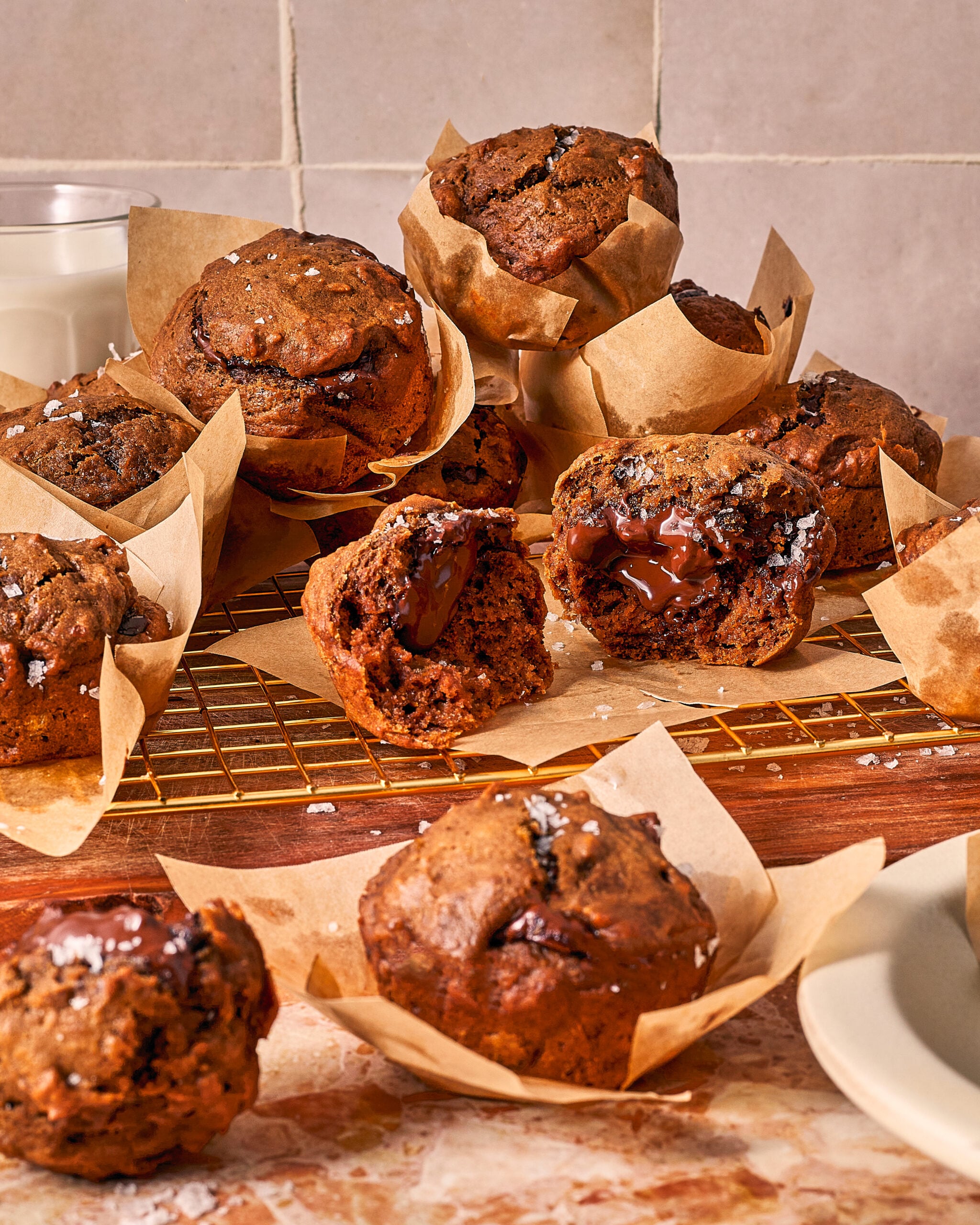 A pile of chocolate muffins in paper liners on a cooling rack, with one muffin broken open to show a melted chocolate center.