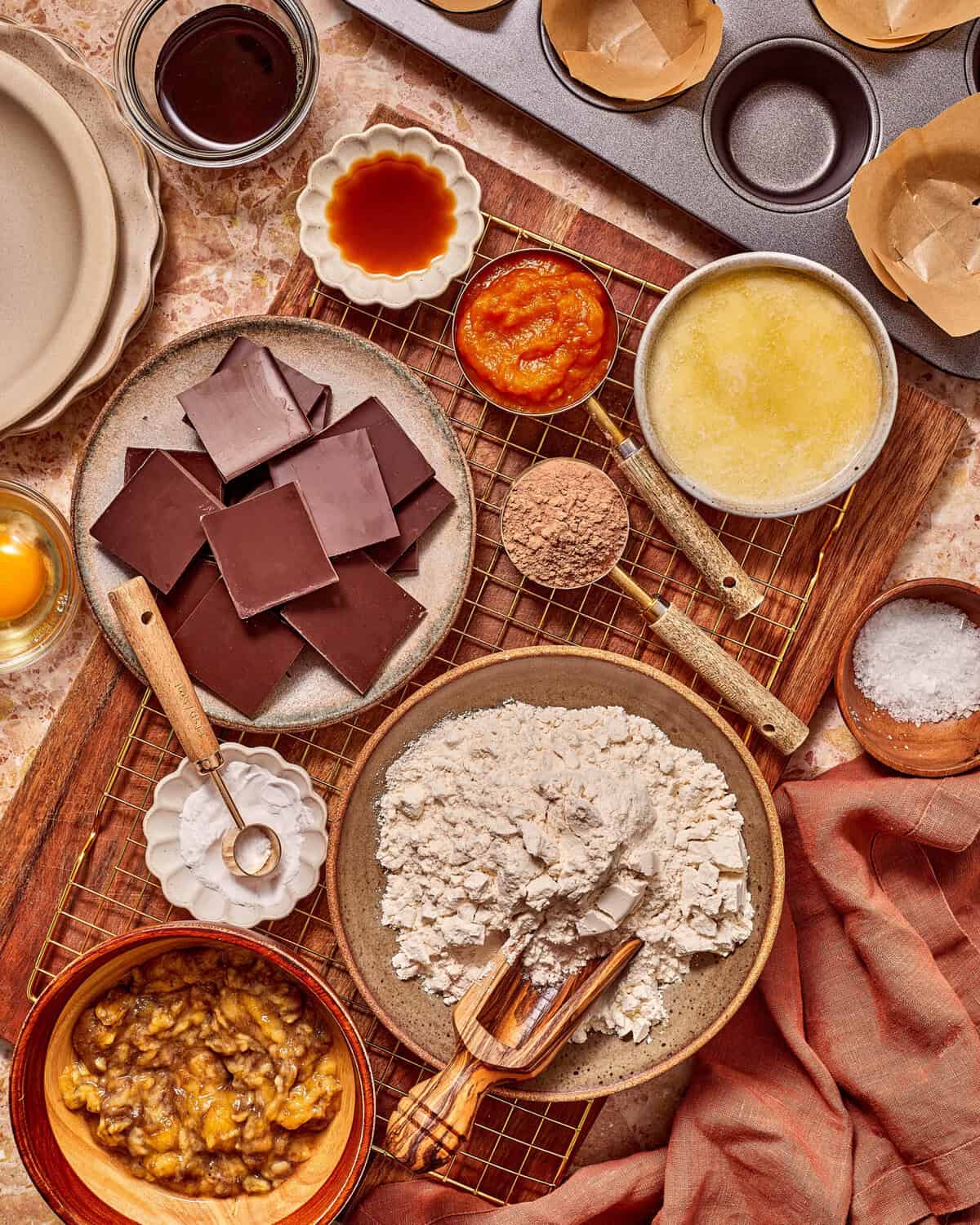 Ingredients for baking arranged on a wooden board, including a plate of chocolate squares, a bowl of flour, mashed bananas, pumpkin puree, melted butter, and various dry seasonings.