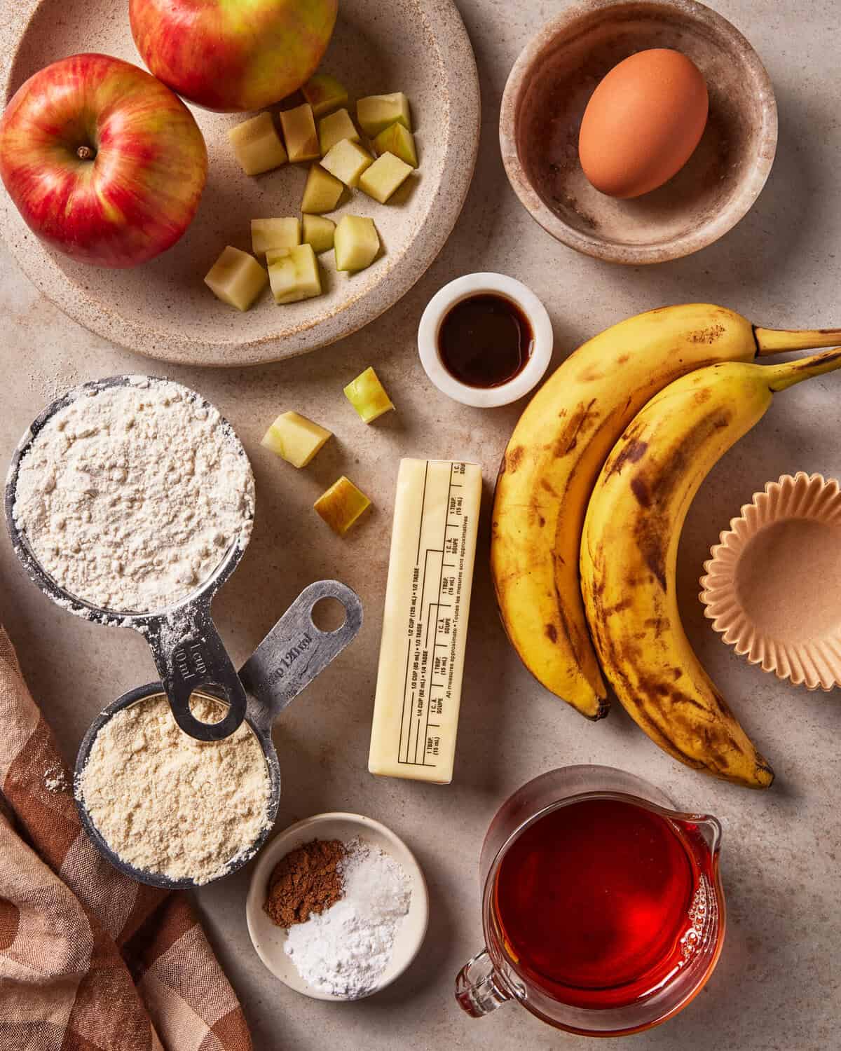 Ingredients for baking arranged on a surface, including two apples, a bowl of diced apple pieces, an egg, two ripe bananas, flour, baking powder, cinnamon, butter, maple syrup, and vanilla extract.