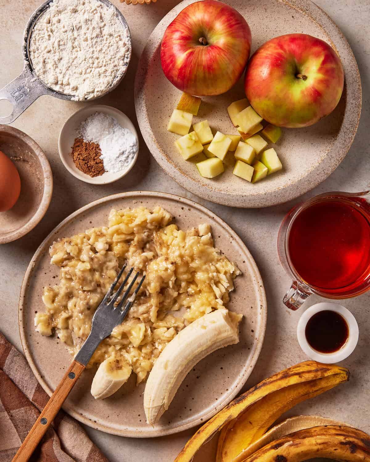 Ingredients for baking including two whole apples, a bowl of diced apples, a bowl of mashed bananas, a measuring cup of flour, an egg, a small bowl of spices, a glass of red liquid, and vanilla extract.