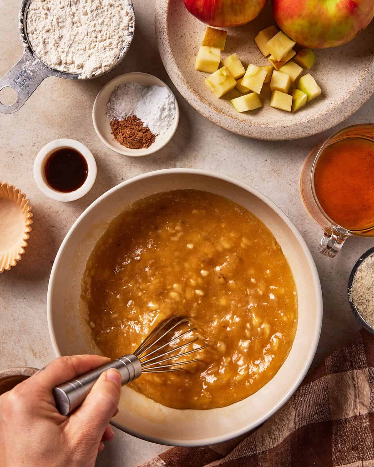 A hand uses a whisk to mix ingredients in a bowl, surrounded by bowls of flour, chopped apples, spices, vanilla, and liquid sweetener.