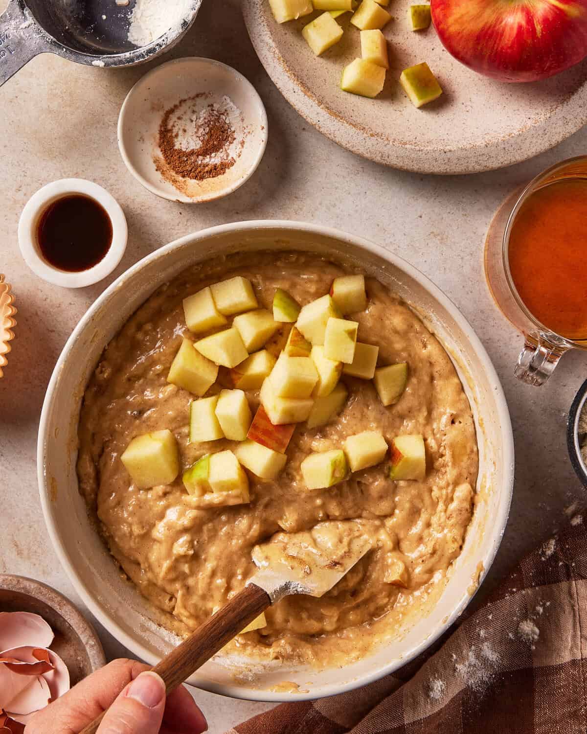 A hand stirs chopped apples into a bowl of thick cake batter with a wooden spatula, surrounded by ingredients including cinnamon, vanilla, and a whole apple.
