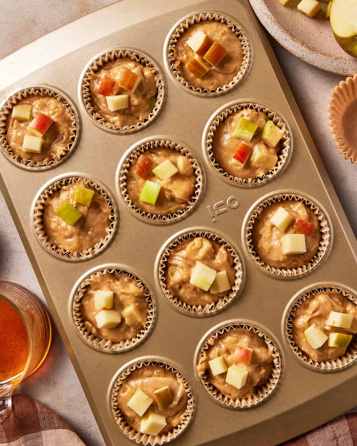 A gold muffin pan filled with raw muffin batter topped with diced apples.