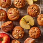 Overhead view of several apple muffins on a wire cooling rack, surrounded by fresh apple halves.
