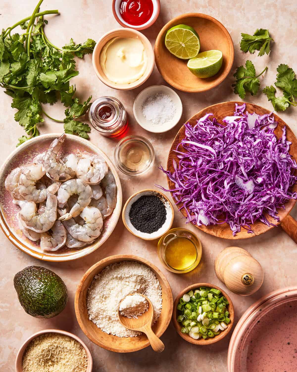 Fresh shrimp, shredded purple cabbage, lime wedges, cilantro, avocado, green onions, sauces, oil, spices, and breadcrumbs arranged in small bowls and plates on a light surface, ready for cooking.