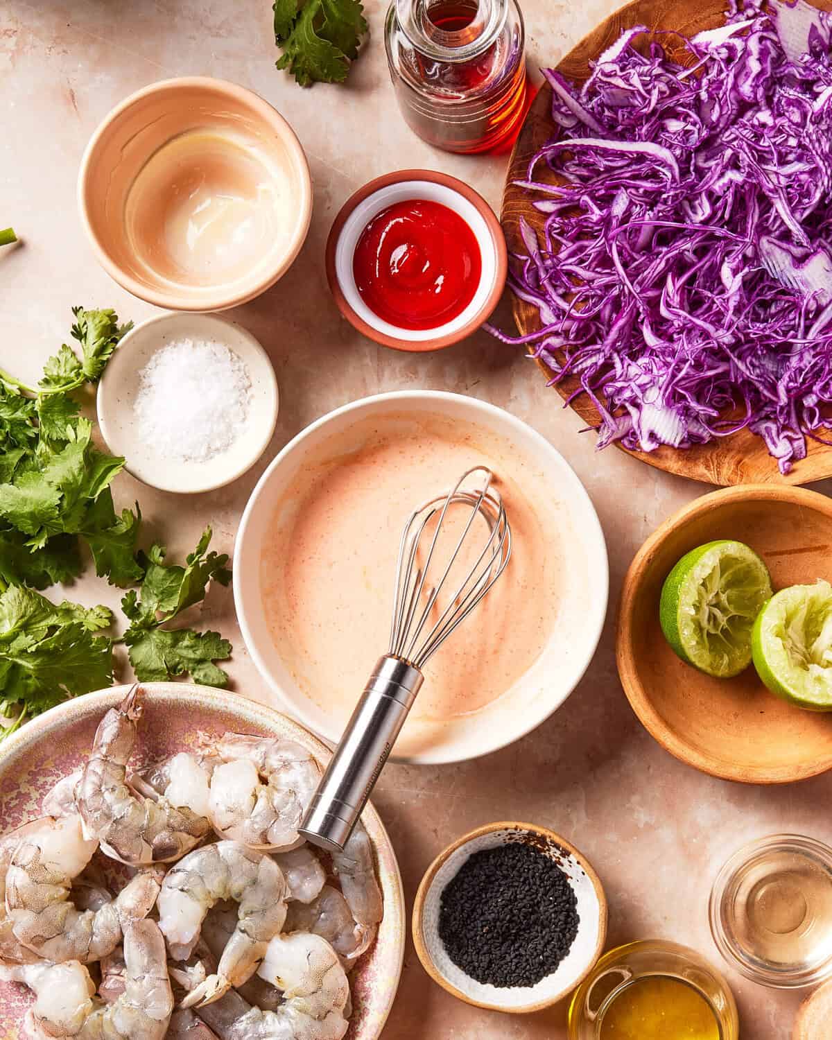 Raw shrimp, shredded purple cabbage, lime halves, cilantro, black sesame seeds, sauces, and a creamy mixture being whisked are arranged on a kitchen counter, suggesting preparation for a seafood dish.