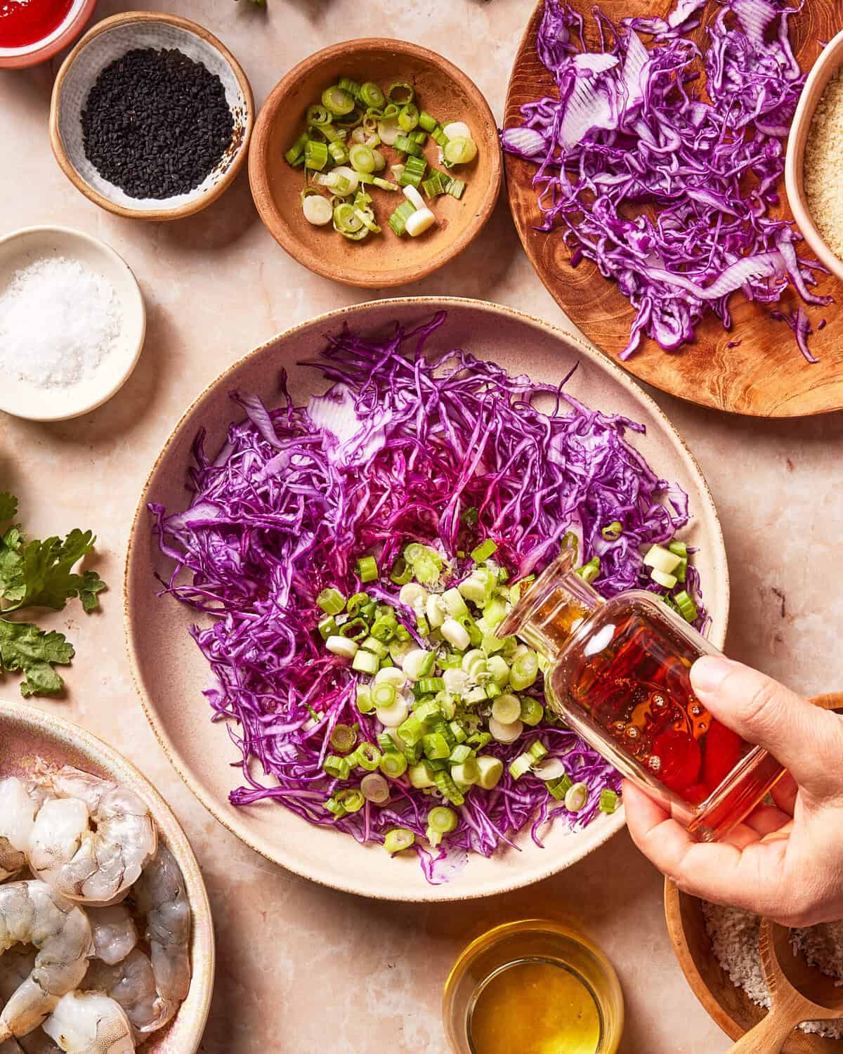 A hand pours oil into a bowl of shredded purple cabbage and chopped green onions, surrounded by sesame seeds, salt, black sesame seeds, shrimp, and other ingredients on a light surface.