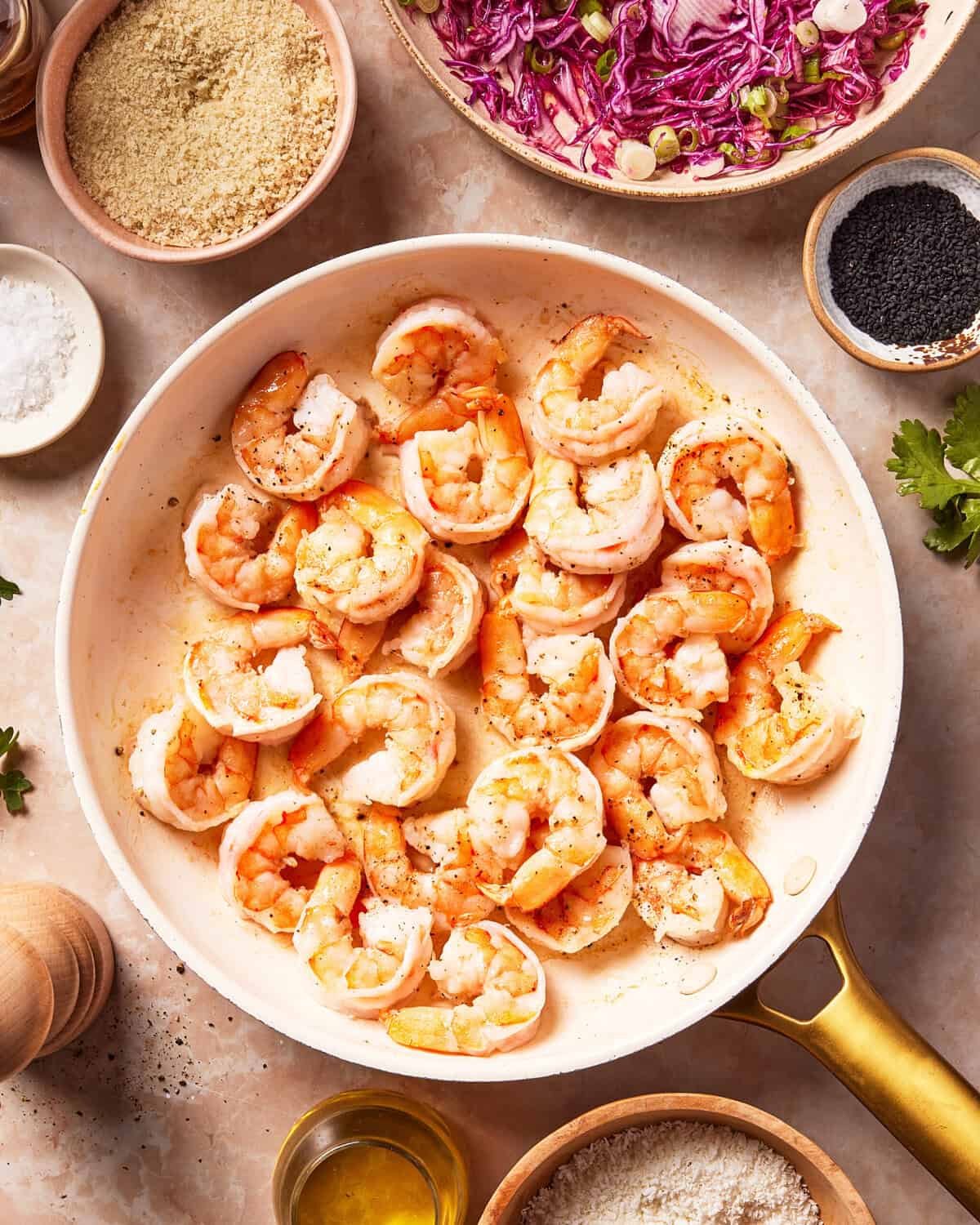 Cooked shrimp in a white skillet surrounded by bowls of breadcrumbs, shredded purple cabbage, black sesame seeds, salt, oil, and fresh cilantro on a light countertop.