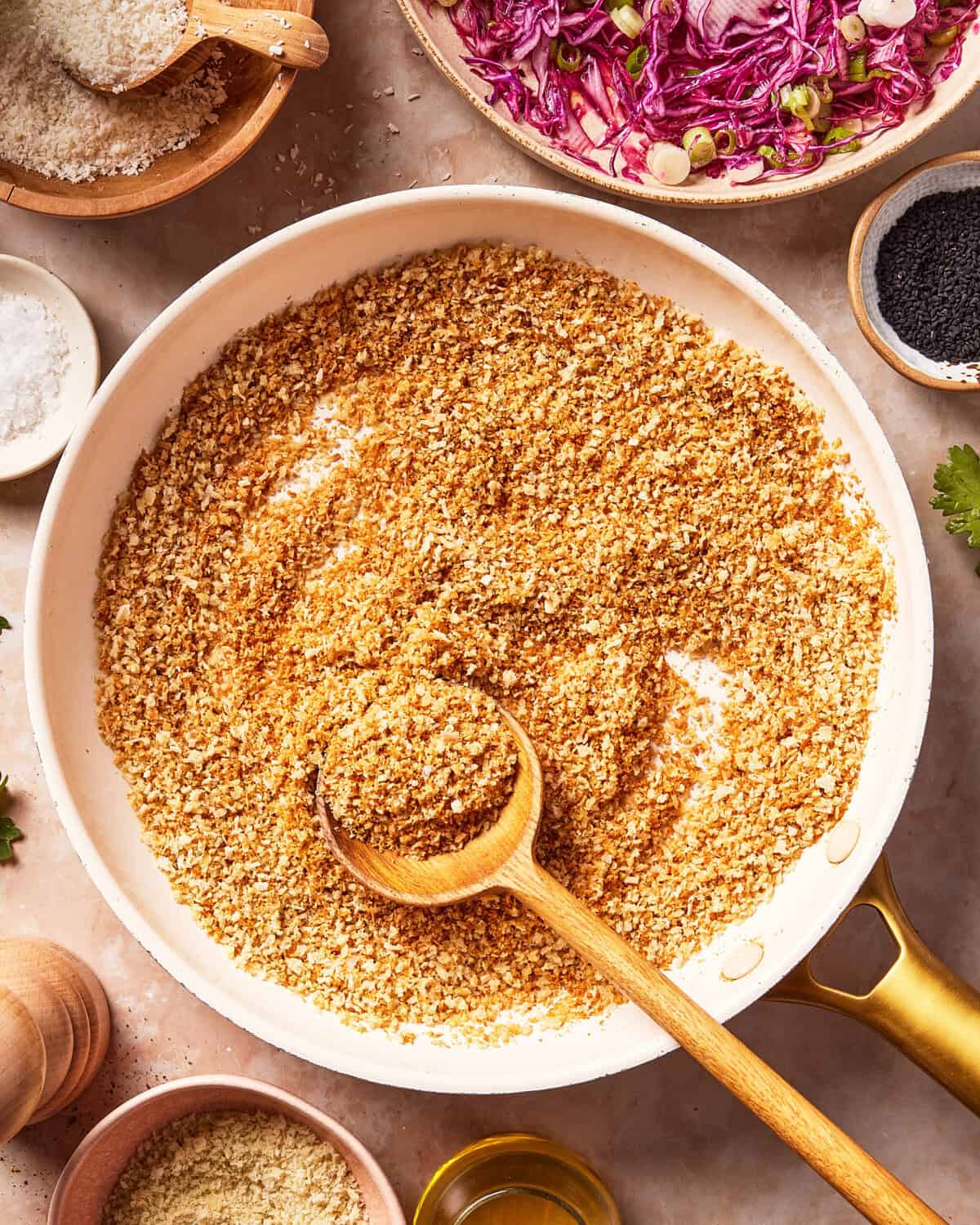 A skillet filled with golden toasted breadcrumbs, a wooden spoon resting in it. Surrounding the skillet are bowls of shredded red cabbage, seeds, grated cheese, and salt, all on a light countertop.